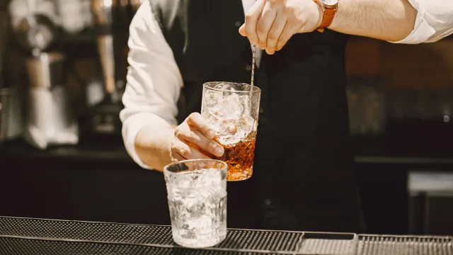 luxury, people, resort - Un bartender professionista in gilet mescola un cocktail con ghiaccio in un bicchiere lavorato al bancone di un bar.