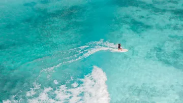 activity, drone-shot, sea - Aerial drone view of a surfer gliding on a surfboard over crystal clear turquoise waters with white ocean spray trails