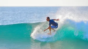 activity, sea - A fit man surfing a vibrant turquoise wave in the ocean, creating a large water spray under a bright sunny sky.