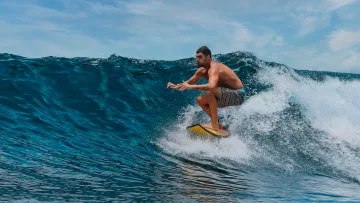 activity, people, sea, sky - A fit shirtless man skillfully riding a powerful blue ocean wave on a yellow surfboard under a bright sunny sky