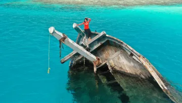 activity, people, sea - A woman posing on the bow of a partially submerged wooden shipwreck in the bright turquoise waters of Vaavu Atoll.