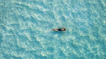 drone-shot, people, relax, sea - Aerial view of a woman in a black swimsuit floating peacefully on the surface of crystal clear turquoise ocean water