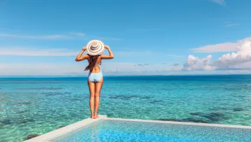 luxury, people, resort, sea - A woman in a striped swimsuit and sun hat stands at the edge of an infinity pool overlooking the blue ocean horizon