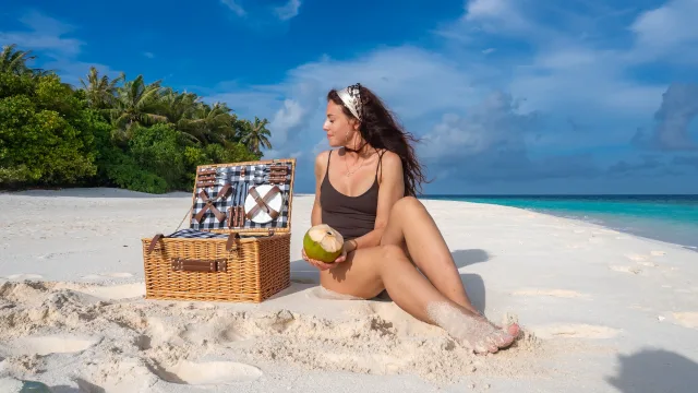 activity, beach, food, luxury, people - A woman in a black swimsuit holding a fresh coconut during a luxury beach picnic with a basket on white sand.