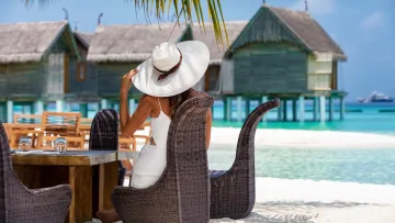 beach, luxury, people, relax, resort - Una donna con cappello da sole e abito bianco siede al ristorante in spiaggia con vista sulle ville acquatiche.
