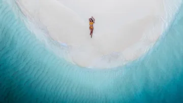 beach, drone-shot, people, relax, sea - Aerial view of a woman in a bikini sunbathing on a white sandy beach surrounded by turquoise ocean waters in paradise.