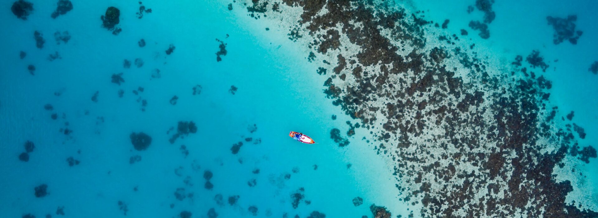 A small boat floating in vibrant turquoise waters next to a dark coral reef, captured from a high aerial perspective.