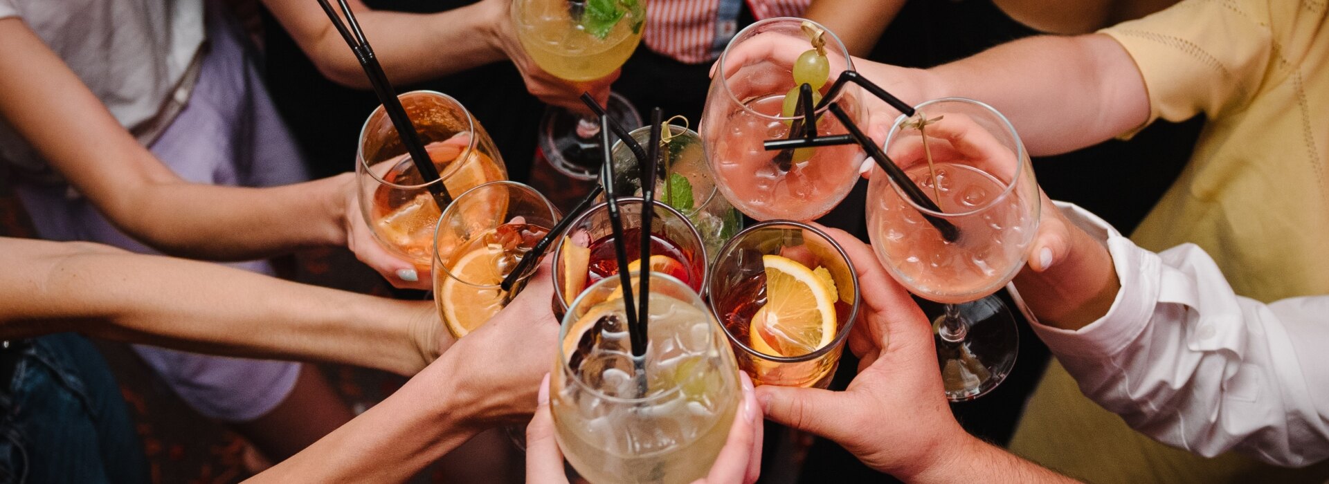 Gruppo di amici che brinda con cocktail colorati decorati con frutta e cannucce durante un aperitivo al tramonto.