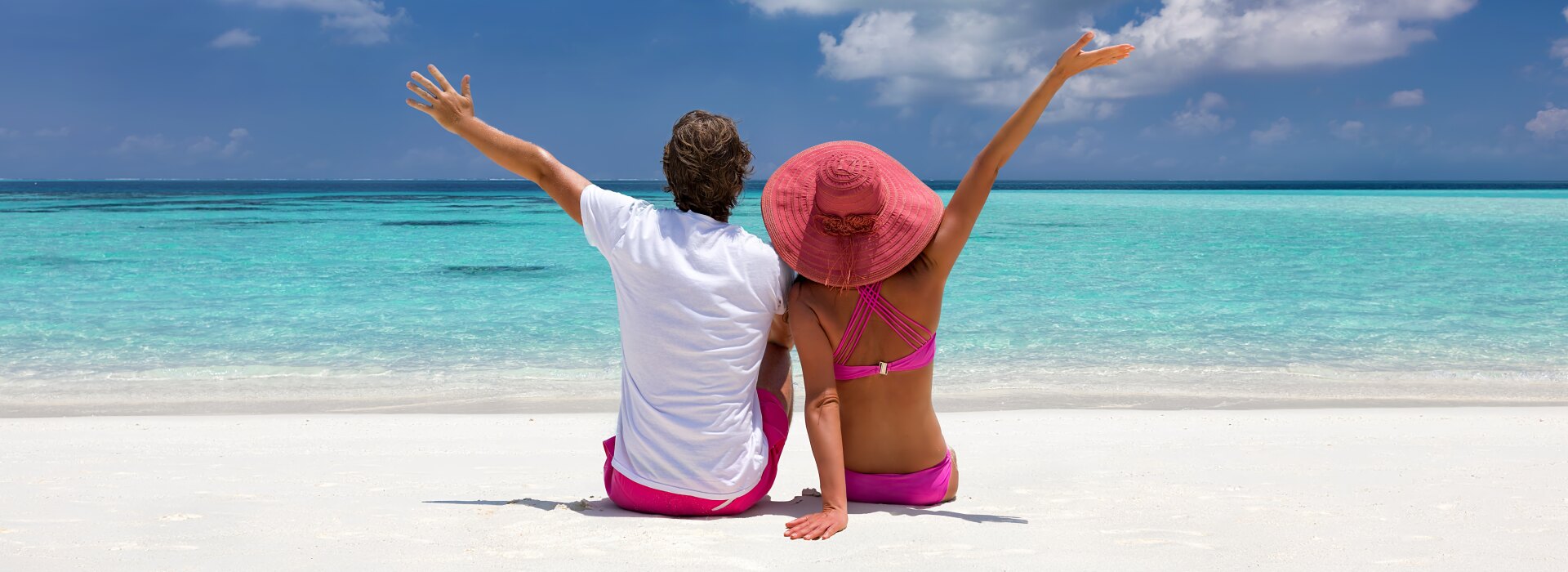 A couple sitting on a tropical beach with white sand and a vast blue sky.