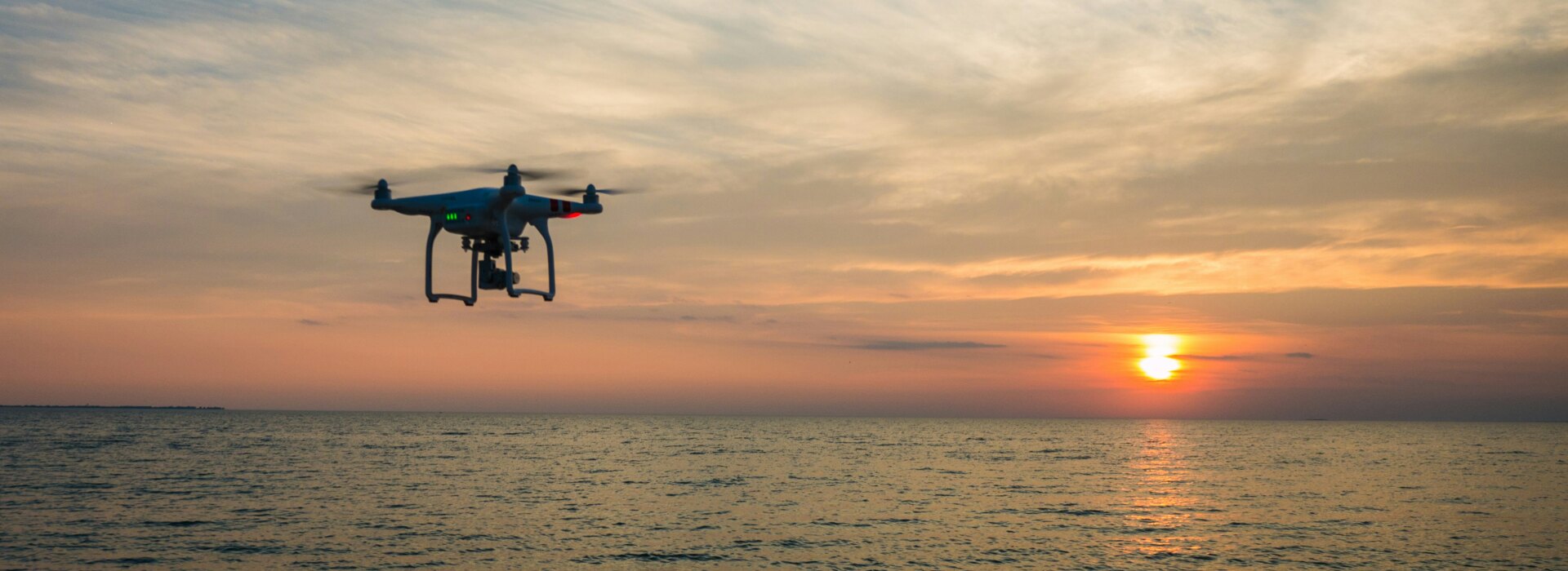 A white quadcopter drone flying over the calm ocean surface during a vibrant golden sunset with clouds above.