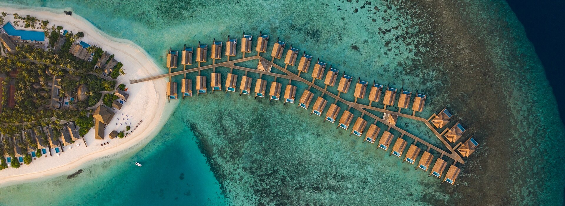 Drone view looking down at a line of luxury overwater villas connected by a wooden pier on a vibrant turquoise reef.