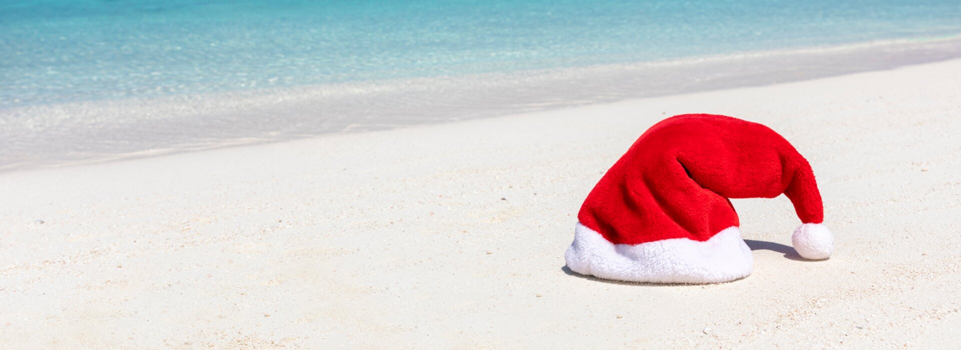 A vibrant red and white Santa Claus hat resting on pristine white sand against a bright turquoise tropical ocean.