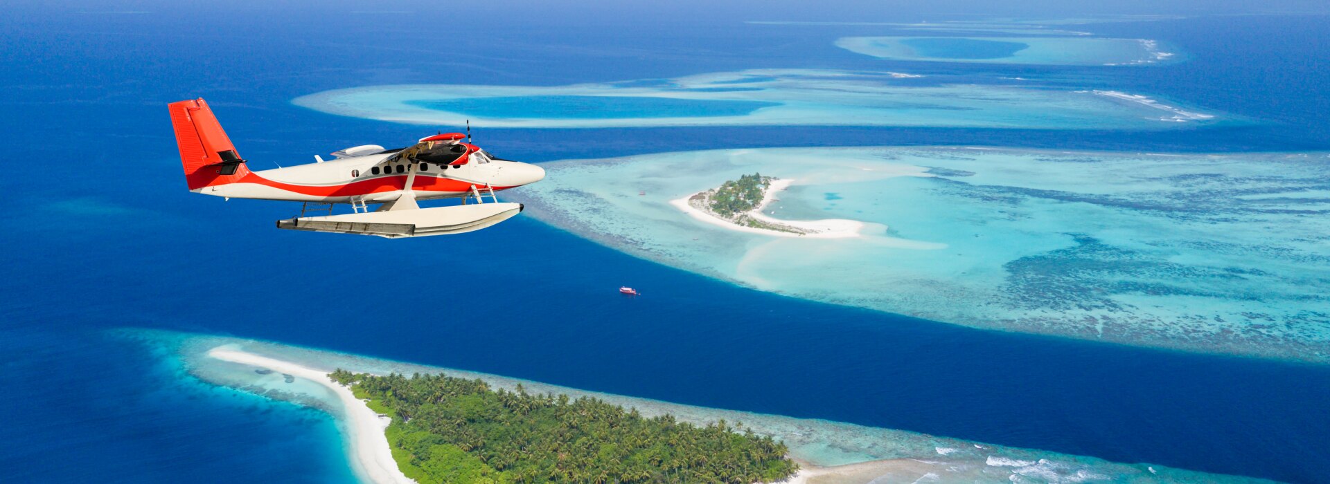A red and white seaplane flying over turquoise waters and lush green tropical islands in the Indian Ocean.