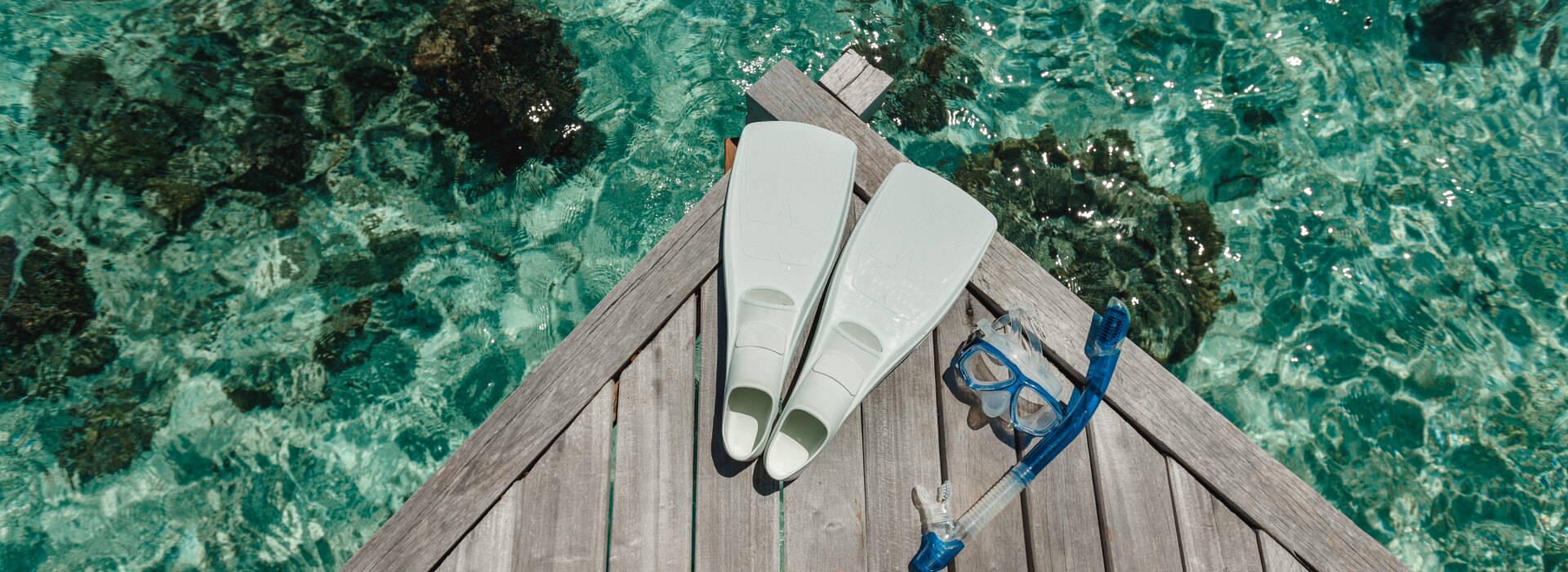 Vista ravvicinata di pinne bianche e maschera blu da snorkeling su un pontile di legno sopra acque tropicali cristalline.
