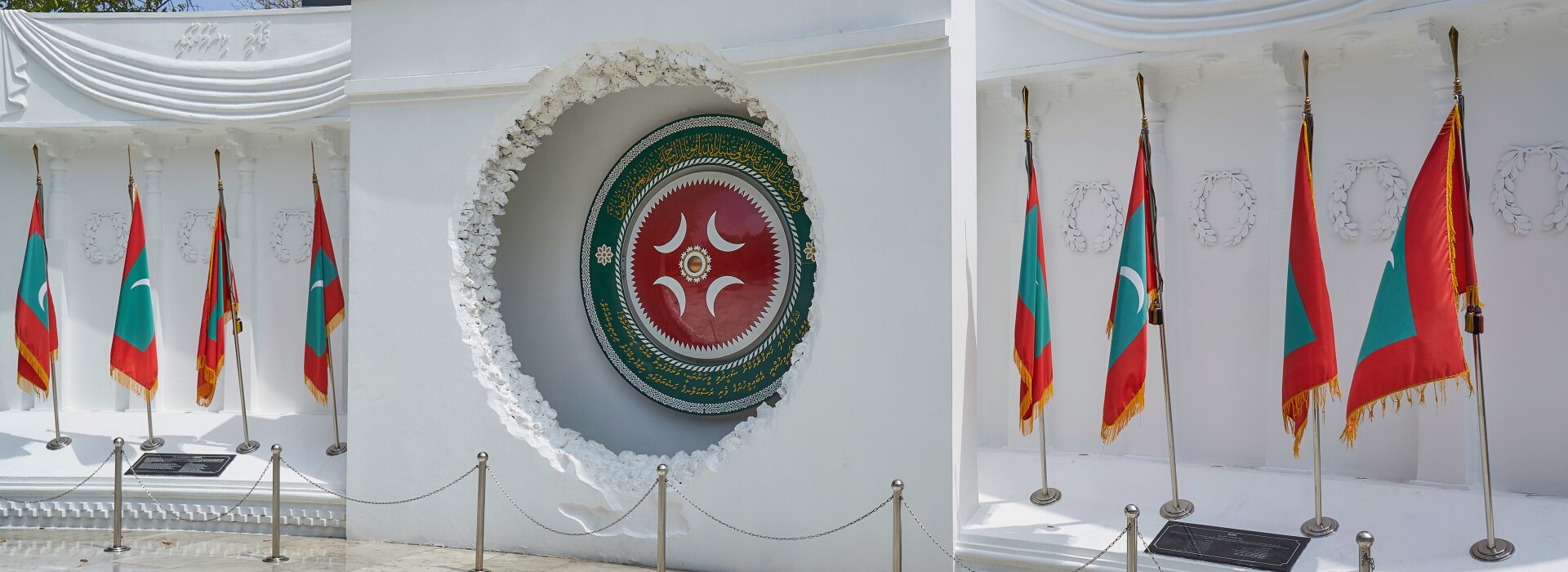 Il Monumento alla Vittoria bianco a Male con l'emblema nazionale e diverse bandiere sotto un cielo limpido.