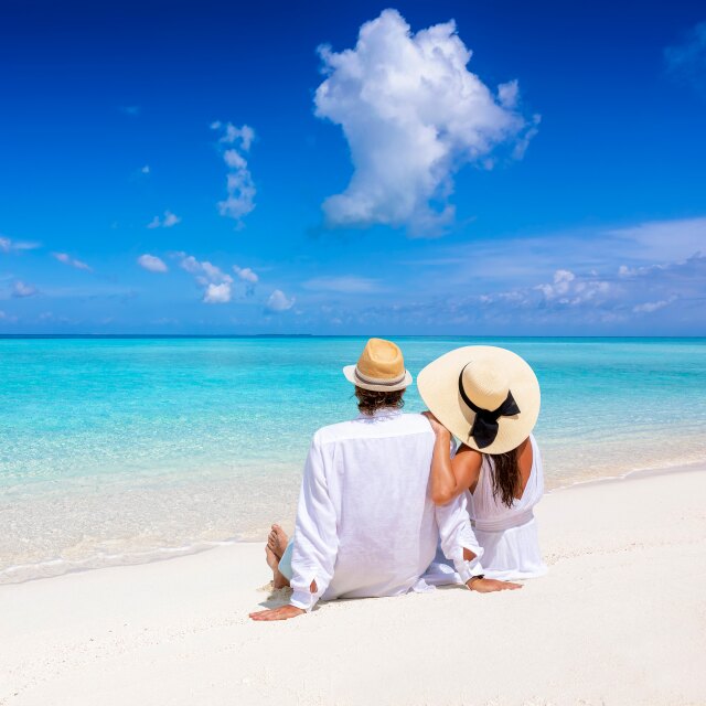 A romantic couple wearing white clothing and hats sitting on white sand, overlooking a bright turquoise ocean.
